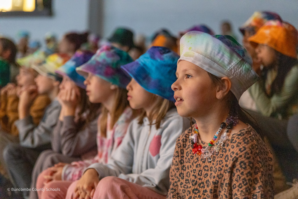 Students sit in a row in the school theater. They are all wearing fishing hats decorated in tie-dye.