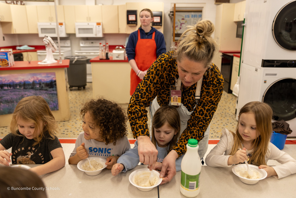 Ms. Rice helps students sitting at a table stir ingredients in their bowls.
