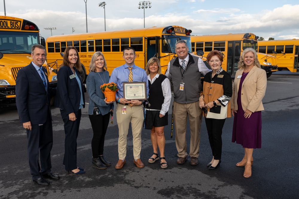 Dr. Jackson, Dr. Johnson, Ms. Harris, Mr. Harbin, Ms. McCurry, Mr. Hough, Ms. Lewis, and Dr. Reed pose for a photo in front of school buses at Enka High. 