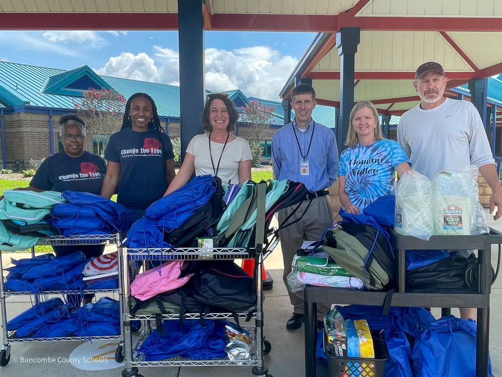 Sydney and her mother, wearing "Change the Type" shirts, stand in a line next to staff at Hominy Valley Elementary. The group is standing behind three carts filled with backpacks and school supplies.