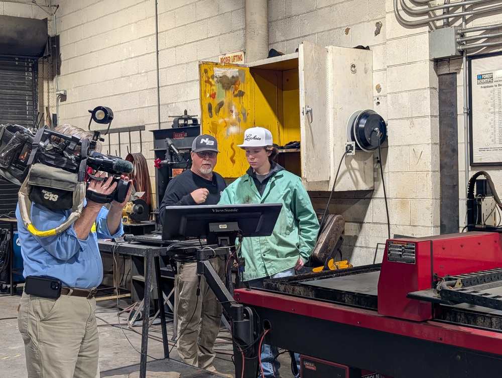 A news photographer points his  camera toward Mr. Fox and a student in the welding class. Mr. Fox is showing the student something on the computer screen.