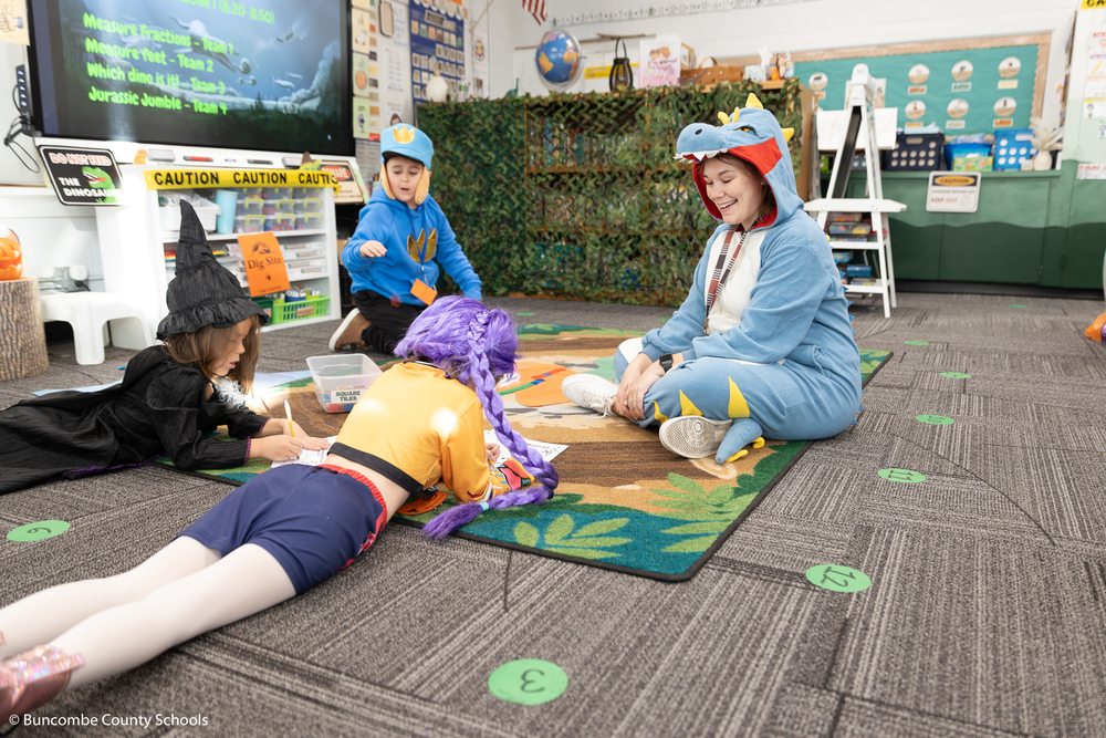 Teacher wearing a dinosaur onesie, sitting on the floor helping students with their dinosaur activity.