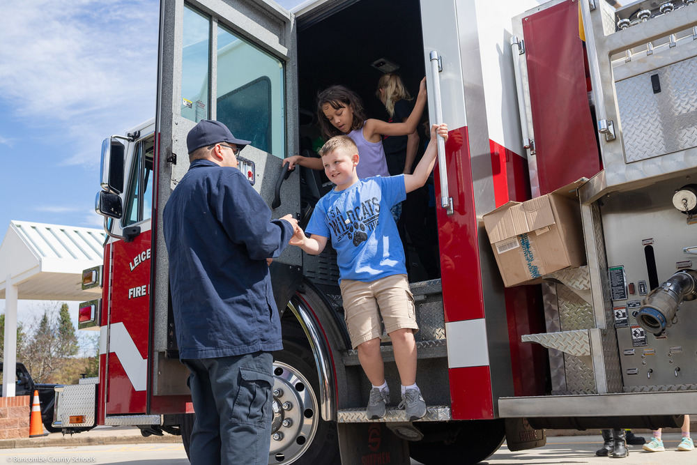 A student steps off of a fire truck while a firefighters hands him a bracelet.