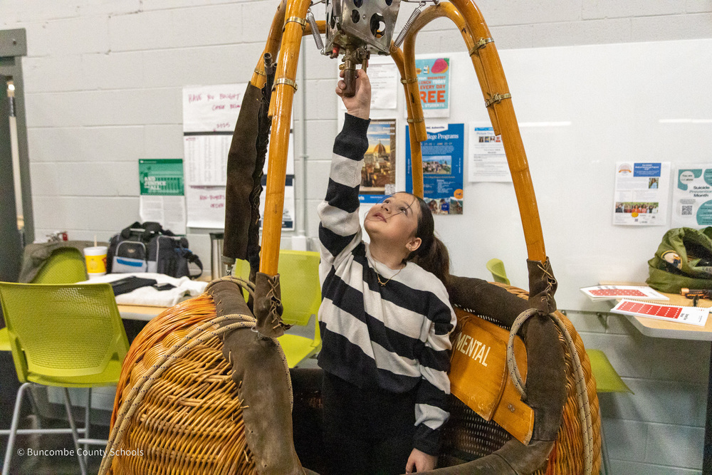 A girl stands in a hot air balloon baaket. She is reaching up to grab a lever.