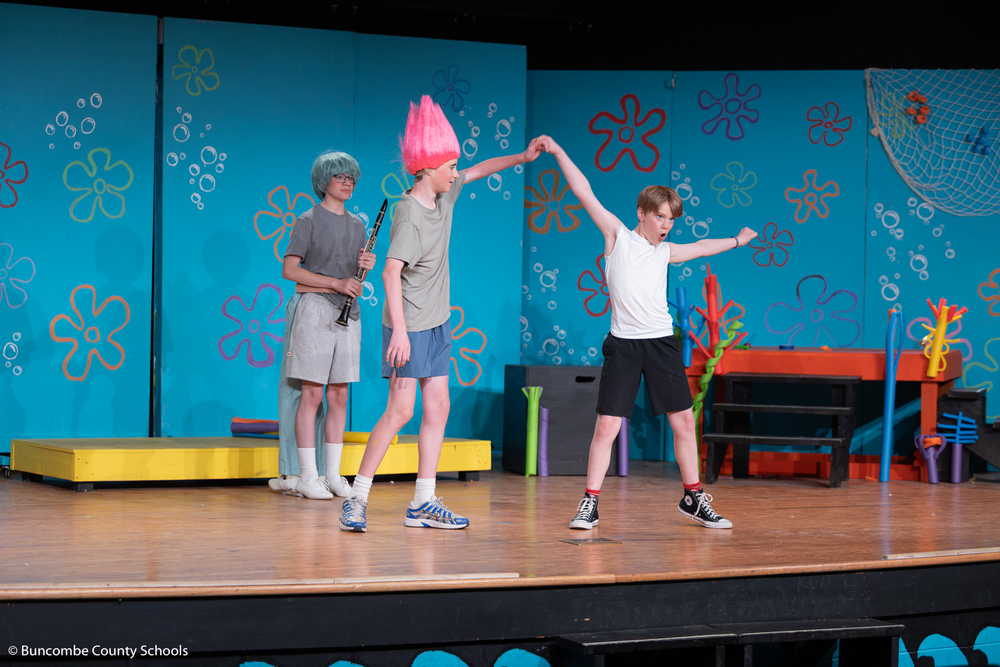 A character in a blue wig with a clarinet and a character in a pink wig stand on stage with a character dancing in a t-shirt on stage at Valley Springs Middle School.