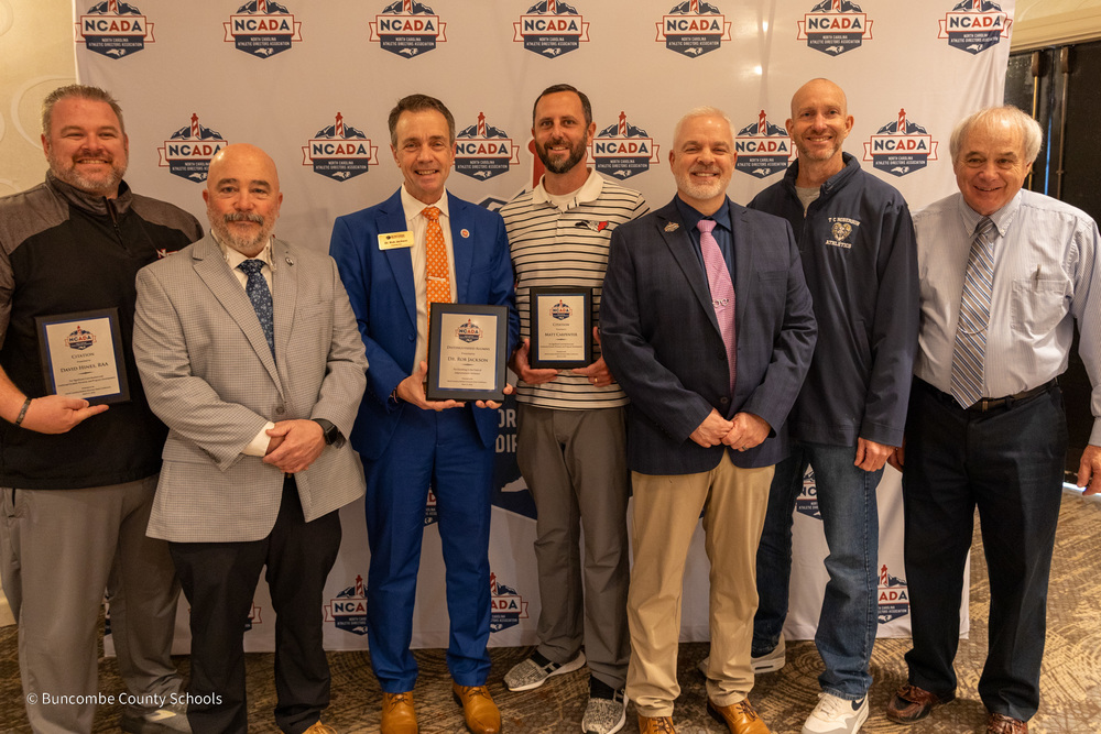 Mr. Hines, Principal Yontz, Dr. Jackson, Mr.  Carpenter, Mr. Carver, Mr. Keen, and Mr. Ball pose for a photo in front of an NCADA banner wall. Mr. Hines, Dr. Jackson, and Mr. Carpenter are holding their plaques.