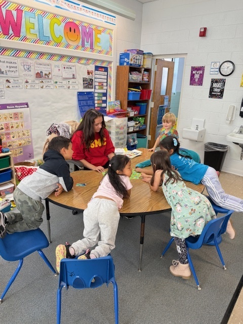 Middle school student sits with five kindergarteners reading a picture book.