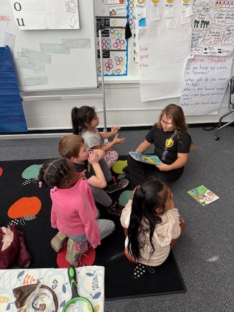 Middle school student sits with four first graders reading a picture book.