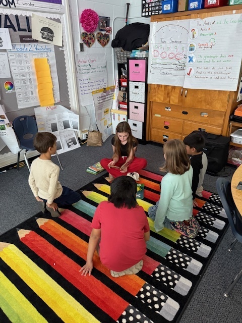 Middle School student sits on floor with four first grade students reading a book. 