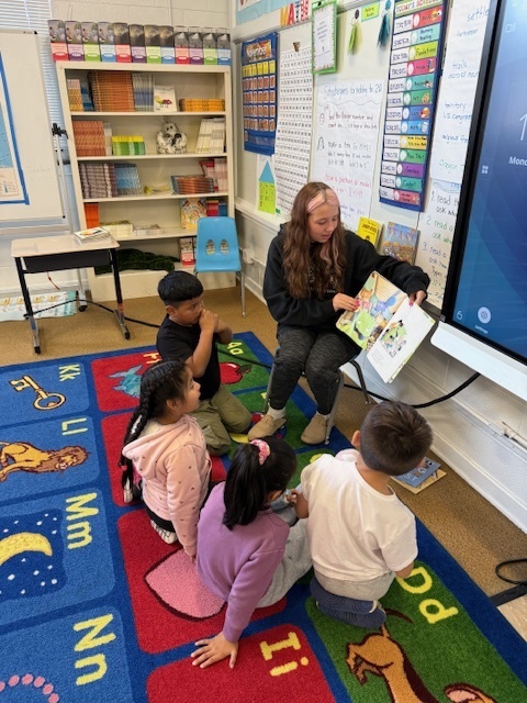 Middle school student holding picture book and reading to four second grade students.