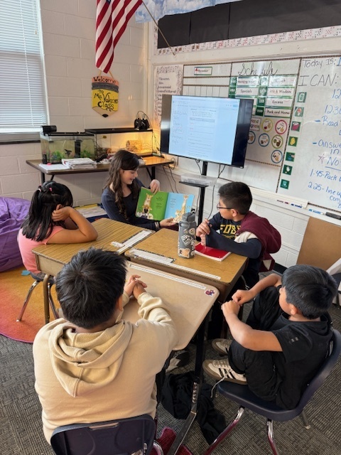 Middle school student sits at desk and reads to four fourth graders.