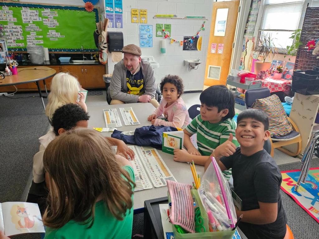 Students sit at table, reading to visiting community member. 