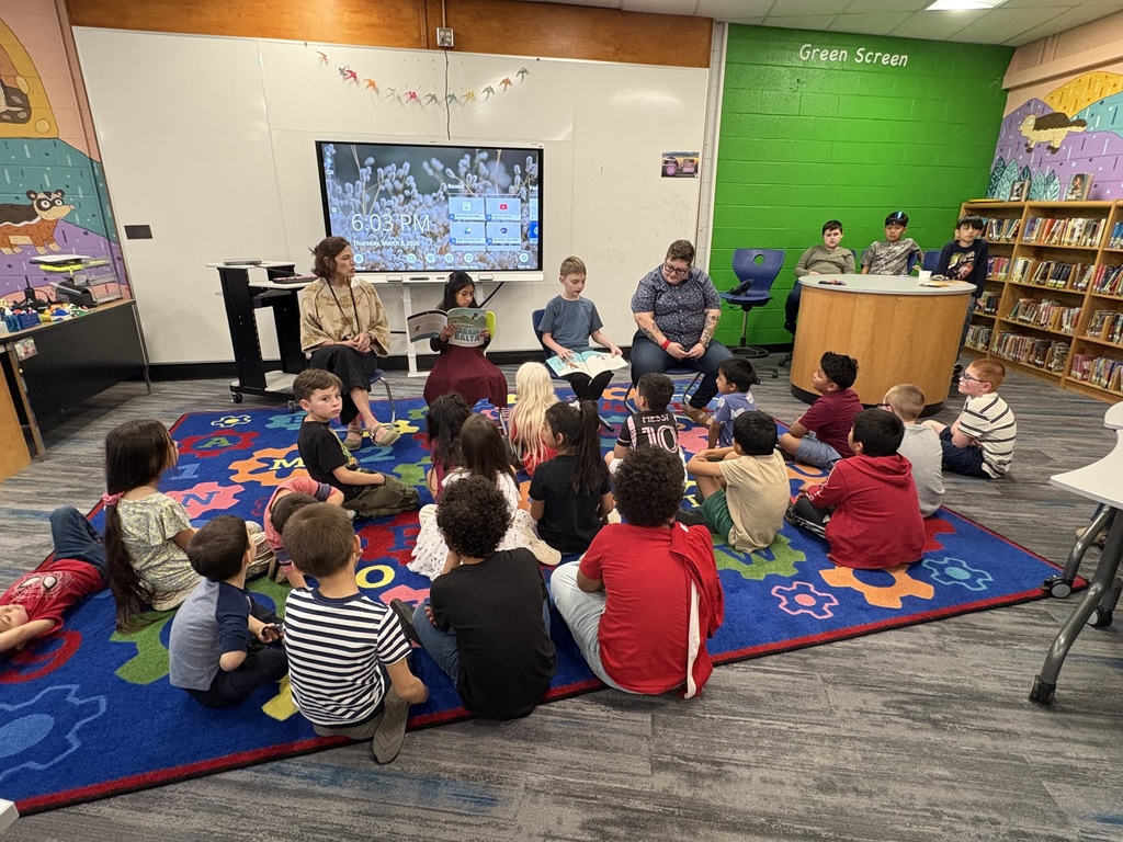 Four people sit in chairs holding books in front of a group of children. 