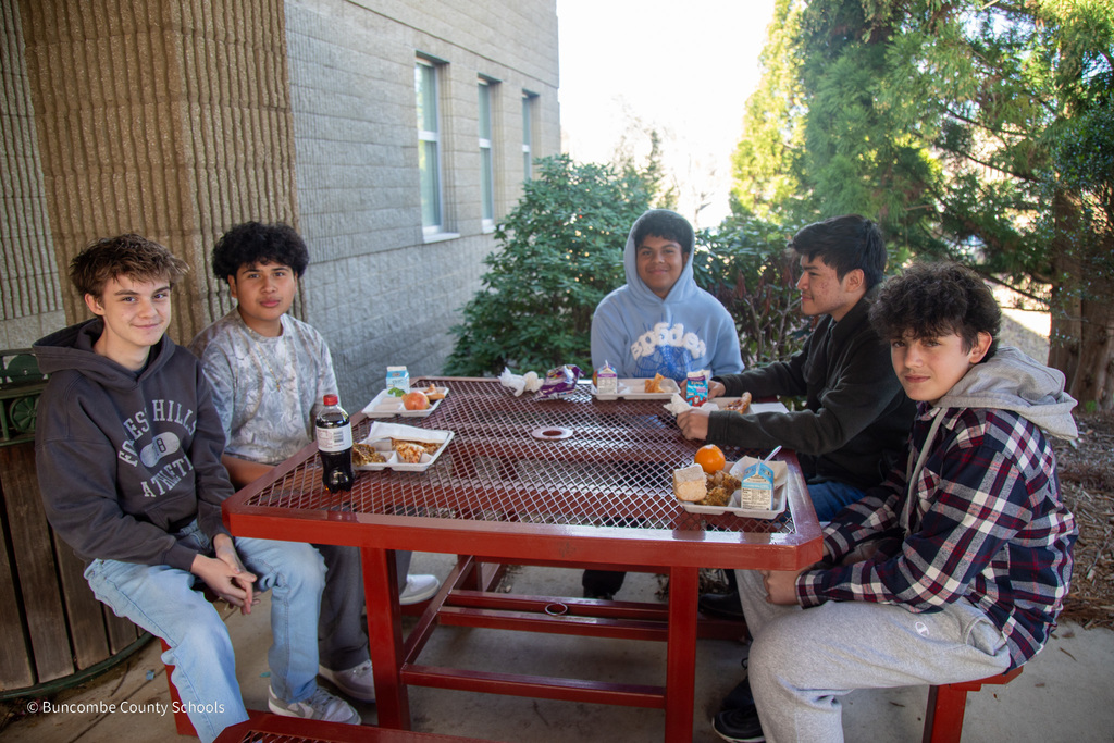 three students sitting outside at a picnic table with lunch trays