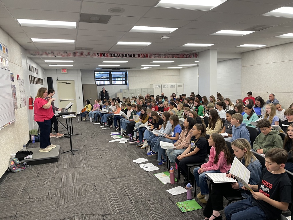 Teacher stands in front of large group of students holding music sheets.