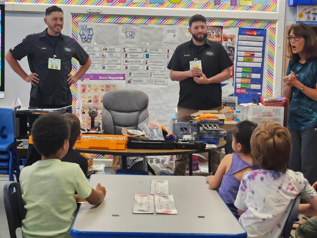 Men stand in front of class smiling while a teacher speaks and four students watch from a table. 