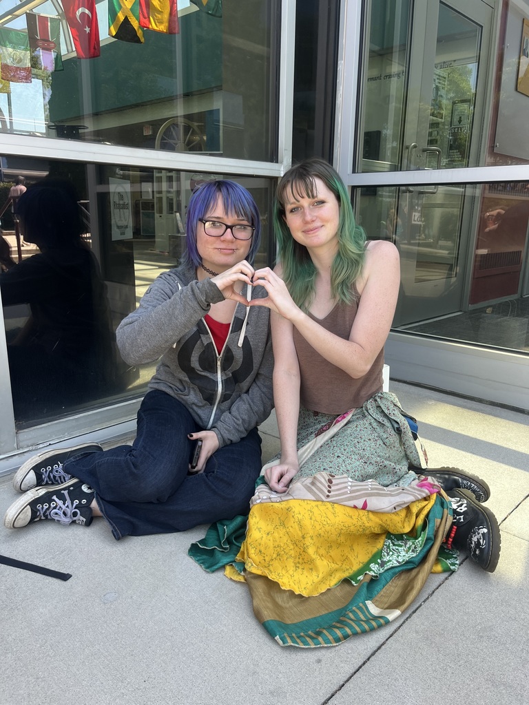 two students sitting outside making a heart with their hands 