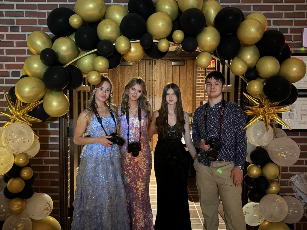 four students holding cameras and standing under a balloon arch at prom