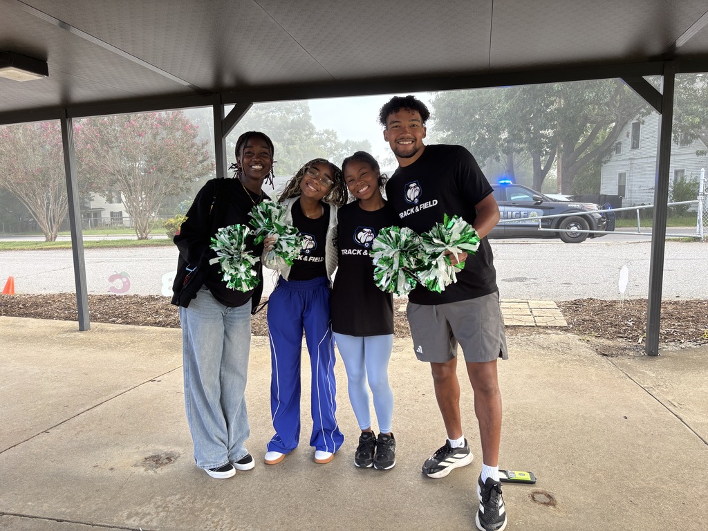 Four UNCA Track and Field Athletes stand with blue and green pom poms to cheer Woodfin students into the building.