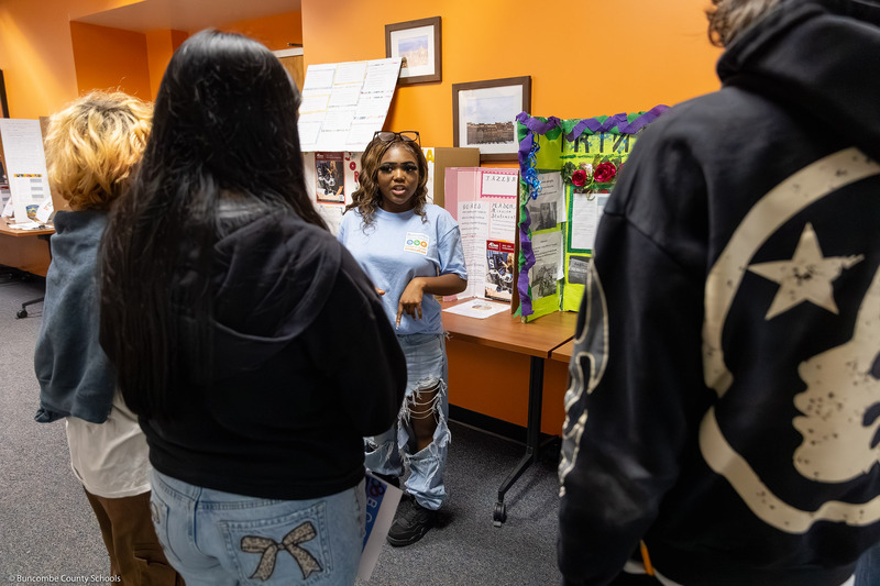 A student speaks to eighth graders in front of her presentation board.