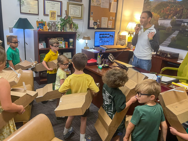 Mr. Alexander using the intercom in his office while students wearing cardboard cars stand in front of his desk.