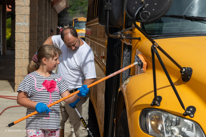 A man and a student use brushes with long handles to wash the exterior of a bus.