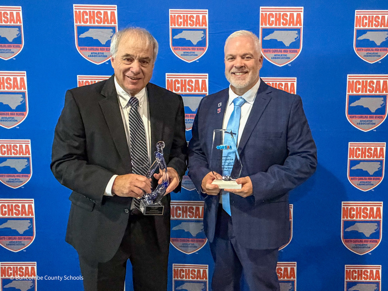 Mr. Ball and Mr. Carver stand in front of the NCHSAA banner wall, each holding their awards.