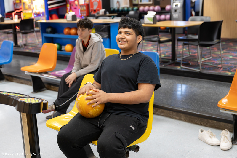 Student holding a bowling ball in his lap as he is sitting and looking at the monitor.