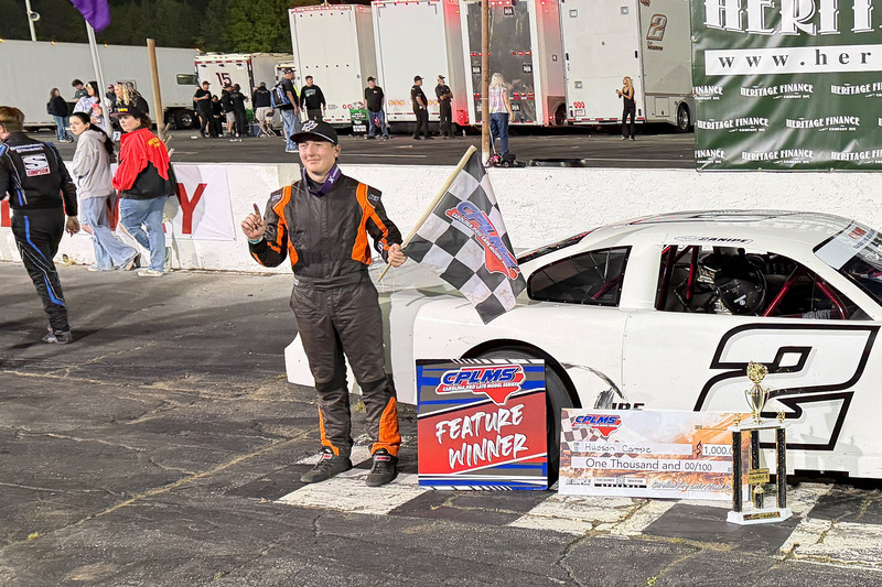 Canpie holds a checkered flag while standing in front of his car.