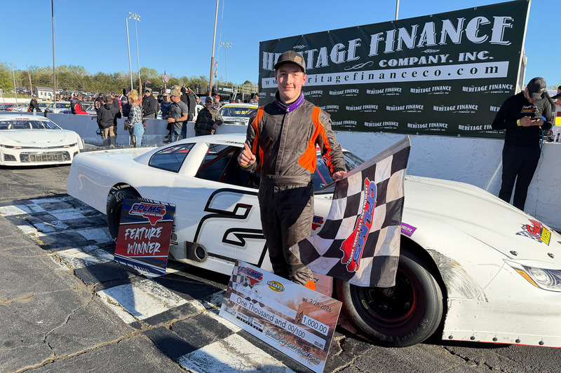 Canpie holds a checkered flag while standing in front of his car.