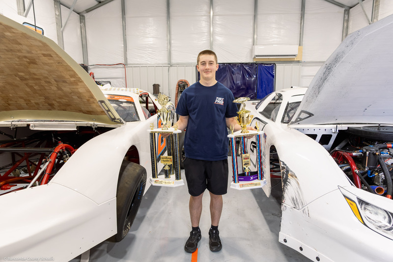 Canipe stands between two racecars while holding two trophies in the workshop.