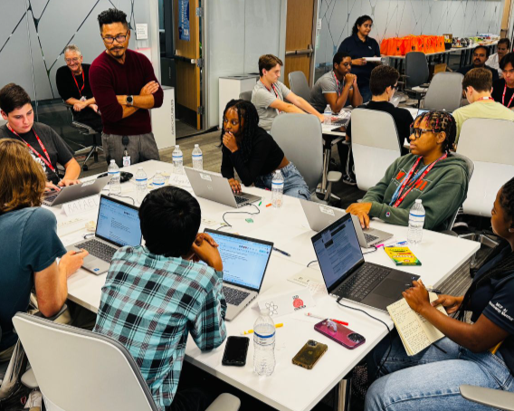 Students with laptops are gathered around a table as an instructor looks at one of the screens.
