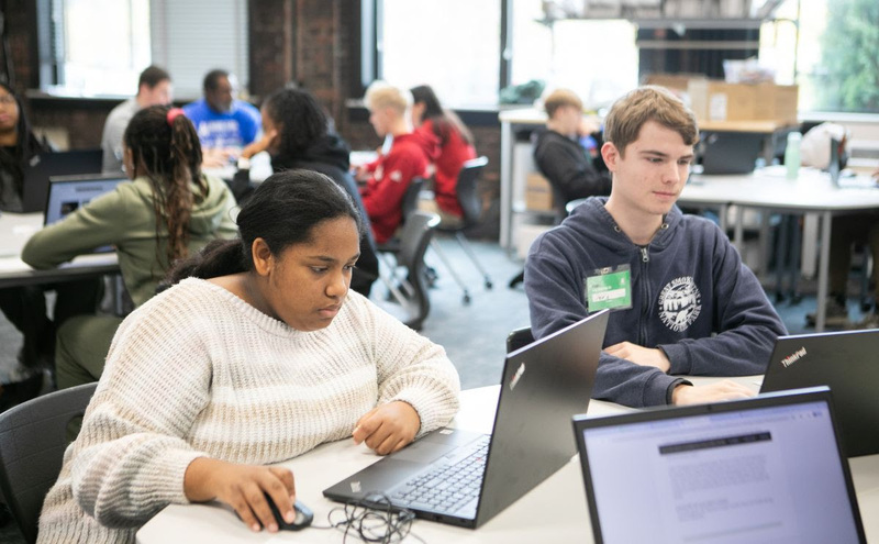 Two students sit at a table working on laptops.