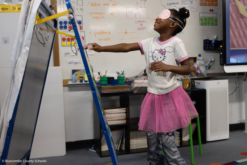 Girl dressed as a princess playing a game of Kiss the frog, where she is wearing a blindfold and trying to put paper lips on a frog poster. 