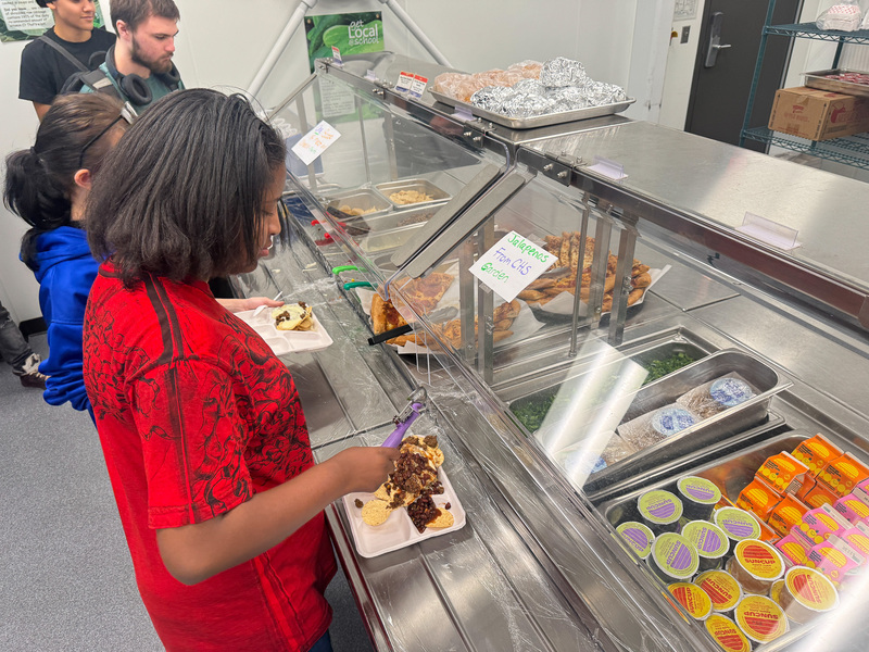 A student going through the school lunch line uses tongs to put jalapenos on her tray.