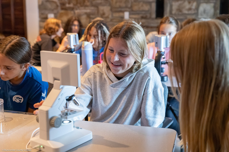 A student smiles while sitting behind a microscope.