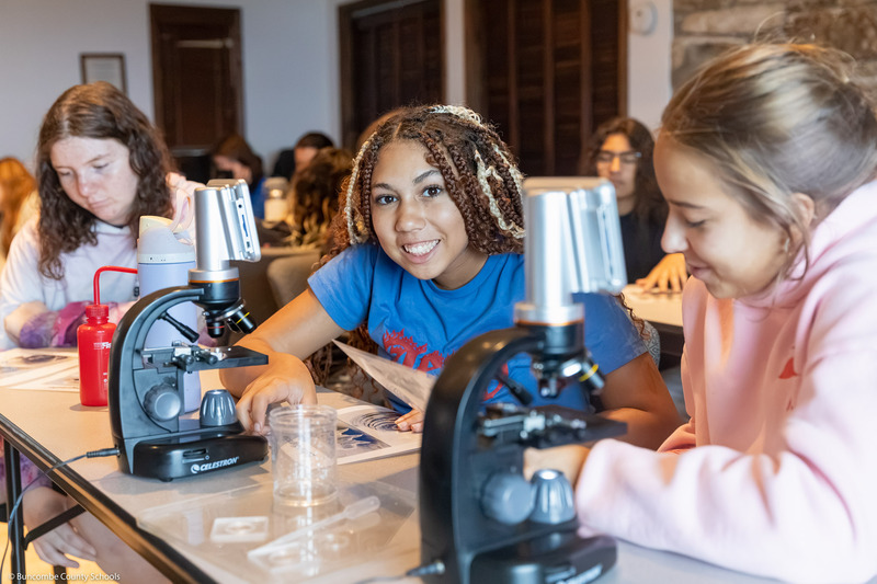 A student smiles while sitting behind a microscope.