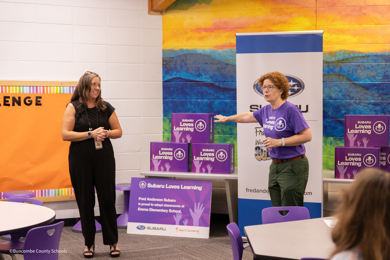 Principal Carla Shipman stands next to a representative from Fred Anderson Subaru. A table of boxes with school supplies is between them.