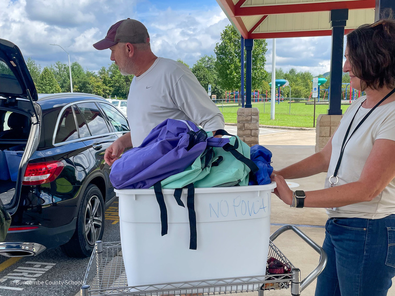 Staff at Hominy Valley stand on the sidewalk near a cart loaded with backpacks