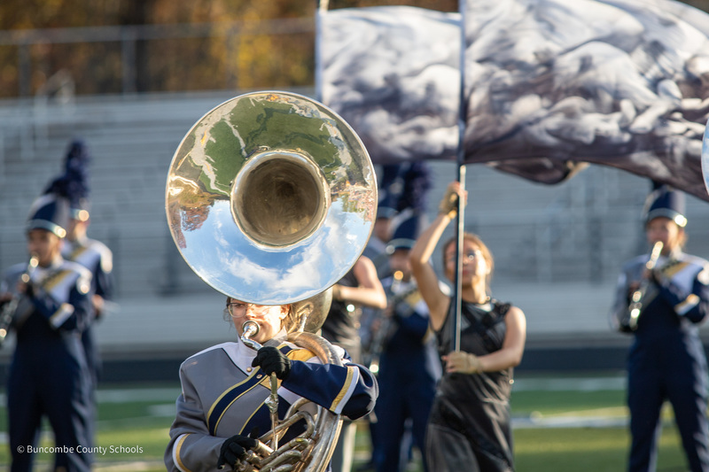 A tuba player from the Roberson band plays her instrument in front of a color guard member holding a black and white flag.