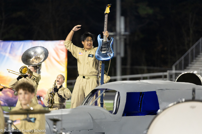 An Enka High band member dressed in a flight suit stands next to an airplane on the field. He is holding his guitar in the air.