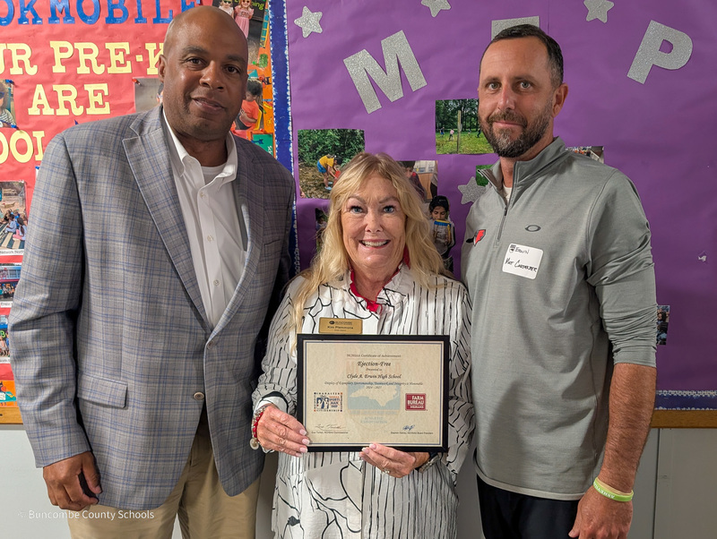 Owen High principal Stanley Wheless, District 6 Board of Education member Kim Plemmons, and Erwin High Athletic Director Matt Carpenter pose for a photo in front of a purple bulletin board. Ms. Plemmons is holding a certificate.