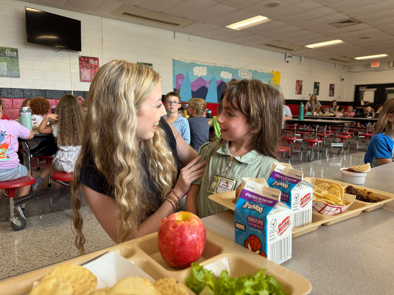 Two sisters in the cafeteria at lunch smiling at each other. 