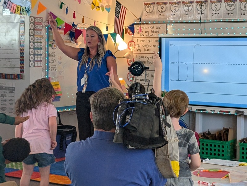 Mrs. Buchanan stands in front of her class pointing to the ceiling. A news photographer from WLOS-TV is pointing his camera in her direction.