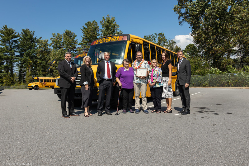 Mr. Cody, Dr. Reed, Mr. Allison, Ms. Franklin, Mr. Honeycutt, Ms. Cheek, Dr. Johnson, and Dr. Jackson pose in front of a school bus. Mr. Honeycutt is holding his plaque.