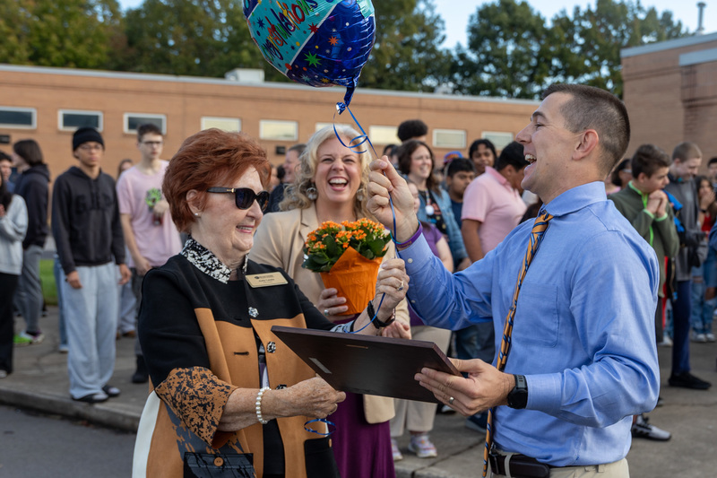 Ms. Lewis presents Mr. Harbin with a balloon as people clap and smile in the background.
