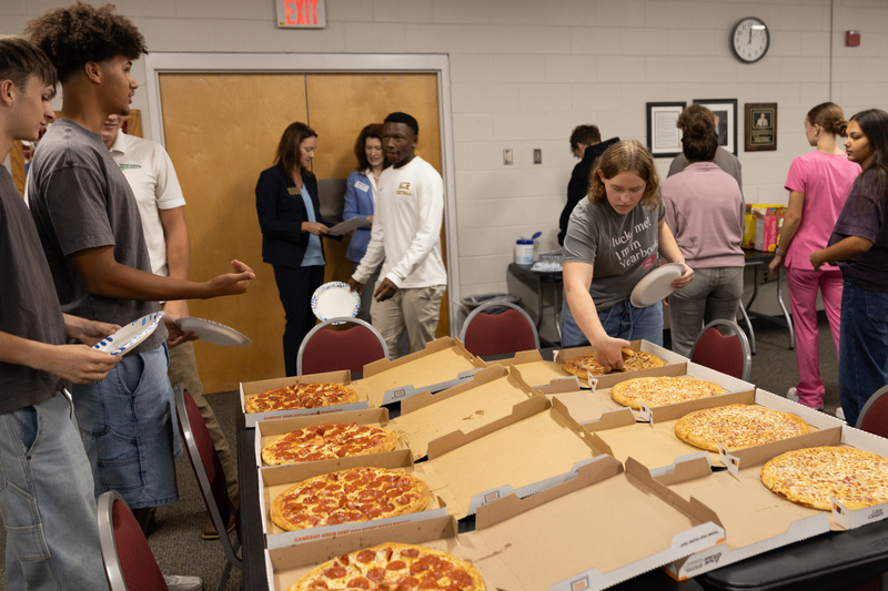 Students stand in front of boxes filled with pizza, taking slices for their lunch