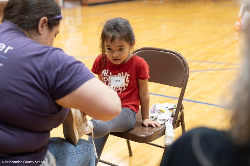 Kindergarten student receiving help from a volunteer with trying on her new shoe. 