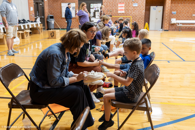 Wide photo showing all the students trying on shoes with the assistance of volunteers. 
