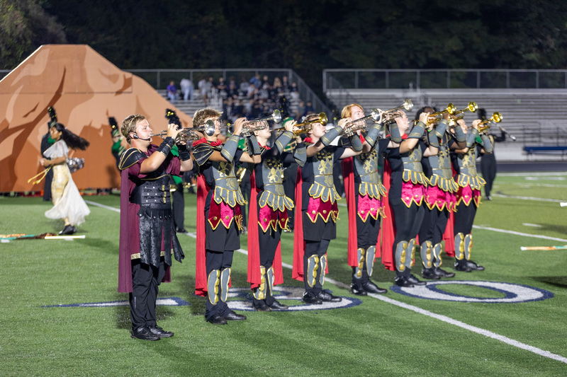 A line of Reynolds band students dressed in Roman soldier costumes playing high brass instruments in front of a wooden volcano.